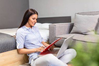 Indoor shot of casually dressed young woman holding papers in her hands, calculating and paying bills at home. Happy beautiful woman sitting on the sofa with laptop calculating home finances