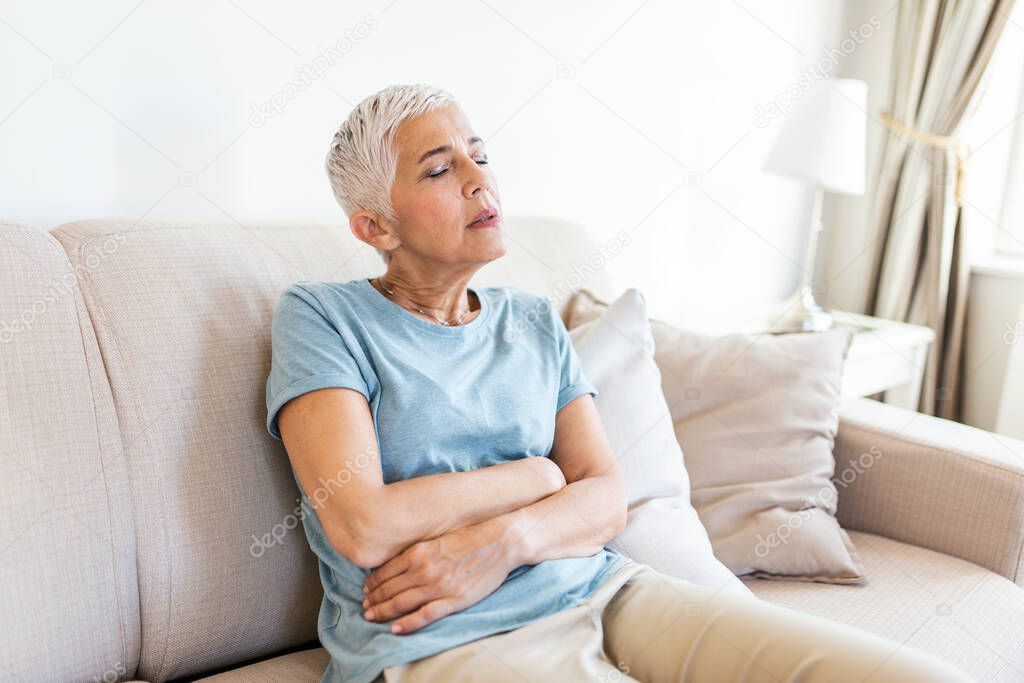 Unhealthy mature woman holding belly, feeling discomfort, health problem concept, unhappy older female sitting on bed, suffering from stomachache, food poisoning, gastritis, abdominal pain, climax