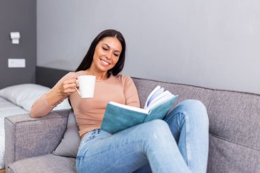 Young beautiful woman lying on sofa and reading book at home. Excellent book. Beautiful young woman reading a book and enjoying her coffee