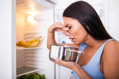 Bad Food In Fridge, young woman in holding her nose because of bad smell from food in refrigerator at home