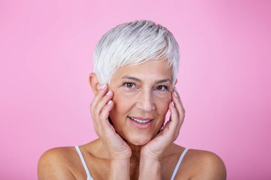 Portrait of beautiful senior woman touching her perfect skin and looking at camera. Closeup face of mature woman with wrinkles massaging face isolated over pink background. Aging process concept.