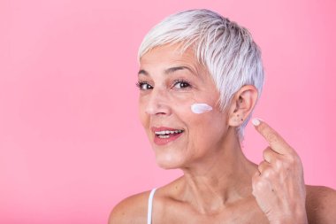 Beautiful mature woman holding jar of moisturizer and looking at camera. Happy senior woman holding a bottle of anti-aging lotion isolated over pink background. Anti aging and beauty treatment.