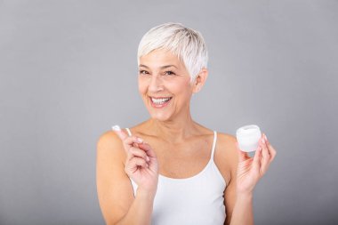Beautiful mature woman holding jar of skin cream for face and body isolated on grey background. Happy senior woman applying anti-aging moisturizer and looking at camera. Beauty anti aging treatment.