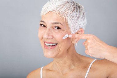 Smiling senior woman applying anti-aging lotion to remove dark circles under eyes. Mature woman using cosmetic cream to hide wrinkles. Lady using day moisturizer to counteract the aging of the skin.