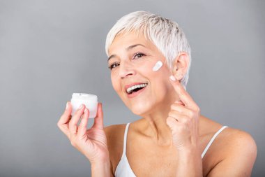 Güzel olgun kadın nemlendirici kavanoz tutarak ve kameraya bakıyor. Gri arka plan üzerinde izole anti aging kremi bir şişe tutan mutlu kadın kıdemli. Anti aging ve güzellik bakımı.