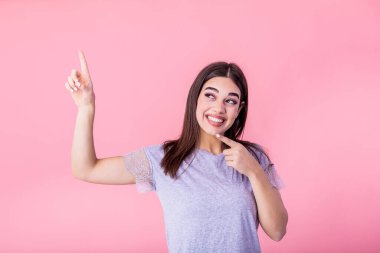 Portrait of brunette woman with long hair in basic t-shirt rejoicing and pointing finger at copyspace isolated over pink background