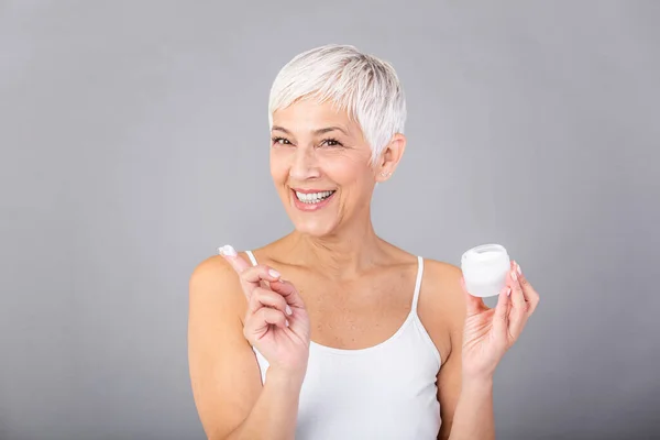 Beautiful mature woman holding jar of skin cream for face and body isolated on grey background. Happy senior woman applying anti-aging moisturizer and looking at camera. Beauty anti aging treatment.