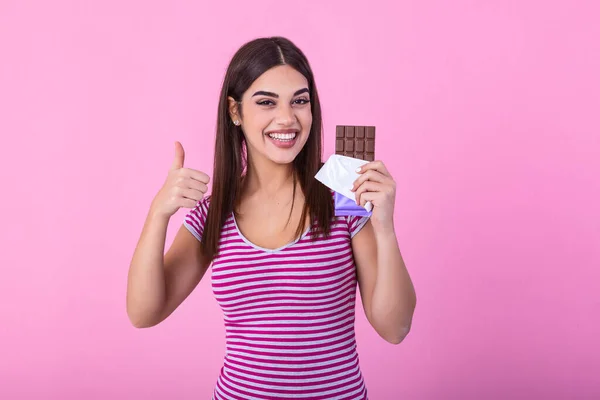 Portrait of a happy young woman with chocolate bar isolated over pink background showing thumbs up. Young woman with natural make up having fun and eating chocolate isolated on pink background