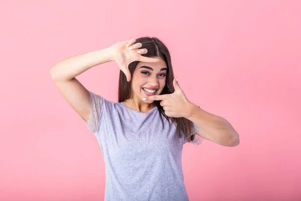Happy young woman making frame gesture with fingers over pink background. Portrait of a happy smiling woman framing her face with her hands isolated