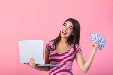 Portrait of an excited satisfied girl holding money banknotes and laptop computer. Happy woman with lucky money. Photo of beautiful woman with laptop, isolated on pink background