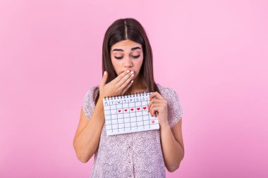 Portrait of a beautiful young girl holding her menstruation calendar and covering hear face isolated over pink background. Period calendar