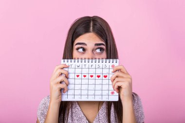 Portrait of a funny young girl in hiding behind a menstrual periods calendar and looking away at copy space isolated over pink background. Female Period calendar