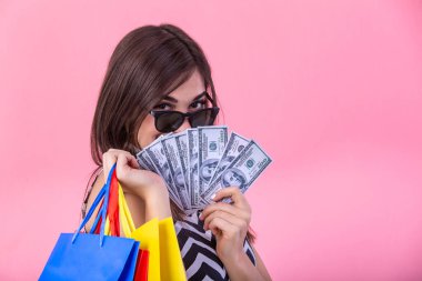 Shopping woman holding shopping bags in one hand and money in other looking at the camera on pink background. Beautiful young shopper smiling happy.
