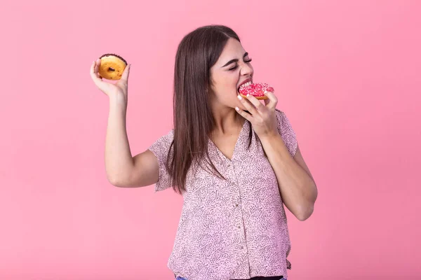 Beauty model girl eating colorful donuts. Funny joyful styled woman choosing sweets on pink background. Diet, dieting concept. Junk food, Slimming, weight loss