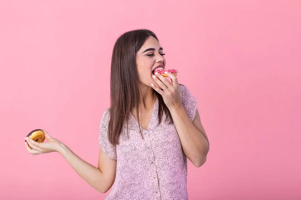 Beauty model girl eating colorful donuts. Funny joyful styled woman choosing sweets on pink background. Diet, dieting concept. Junk food, Slimming, weight loss