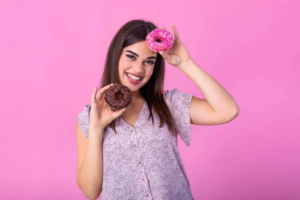 Stylish girl with long hair positively poses, holding fresh pink and chocolate donuts with powder ready to enjoy sweets. Portrait of attractive young woman in retro shirt having fun with sweet-stuff