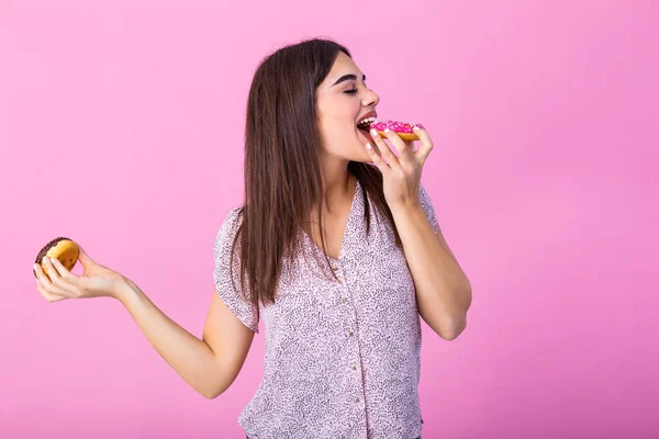 Beauty model girl eating colorful donuts. Funny joyful styled woman choosing sweets on pink background. Diet, dieting concept. Junk food, Slimming, weight loss