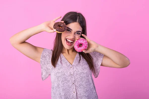 Stylish girl with long hair positively poses, holding fresh pink and chocolate donuts with powder ready to enjoy sweets. Portrait of attractive young woman in retro shirt having fun with sweet-stuff