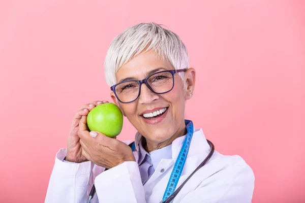 Smiling mature nutritionist in her office, she is holding a green apple and showing healthy vegetables and fruits, healthcare and diet concept