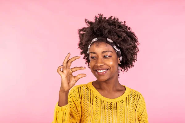 Size matters. Pleased African American young woman demonstrates very tiny object, smiles positively, wears casual sweater, poses against pink background, shapes small thing. Body language concept