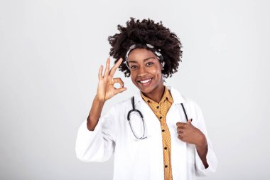 Mutlu Afrikalı kadın doktor beyaz laboratuvar önlüğü giyiyor, steteskop kameraya bakıyor ve tamam işareti gösteriyor. Gülümseyen Afro-Amerikan kadın profesyonel terapist doktor portresi.