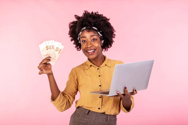 Portrait of excited African American woman holding money banknotes with laptop computer isolated over pink background. Portrait of a cheerful young woman holding money banknotes and laptop computer