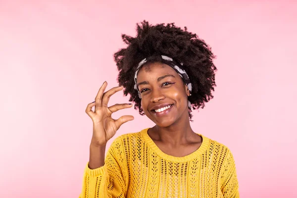 Positive cheerful dark skinned curly woman shapes something very little with fingers, demonstrates small decreased price or salary, gestures not big object, smiles toothily