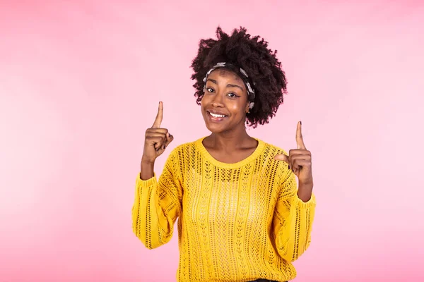 Glad pleased African American woman keeps hands up and points fingers above. Happy dark skinned lady draws your attention to copy space, poses against pink background