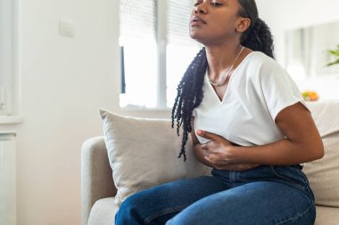 African American Woman in painful expression holding hands against belly suffering menstrual period pain, lying sad on home bed, having tummy cramp in female health concept