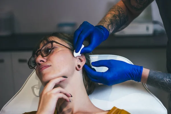 若い女性は彼女の耳をピアス取得 滅菌医療機器やラテックス手袋でピアスのプロセスを示す男 ボディピアス手順 — ストック写真