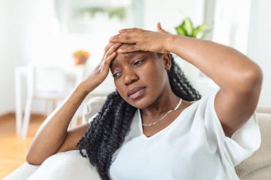 Portrait of a young black girl sitting on the couch at home with a headache and back pain. Beautiful woman suffering from chronic daily headaches. Sad woman holding her head because sinus pain