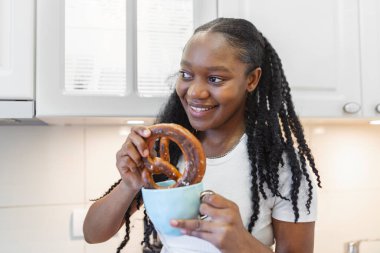 Genç Afrikalı Amerikalı kadın mutfak masasında oturuyor ve bir fincan solucan sütüyle krakerinin tadını çıkarıyor.
