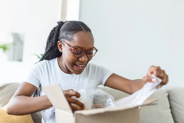 African American woman received ordered goods online via internet website sitting on couch at home enjoy moment unpacking cardboard box parcel feels happy, commerce postal delivery, easy fast service