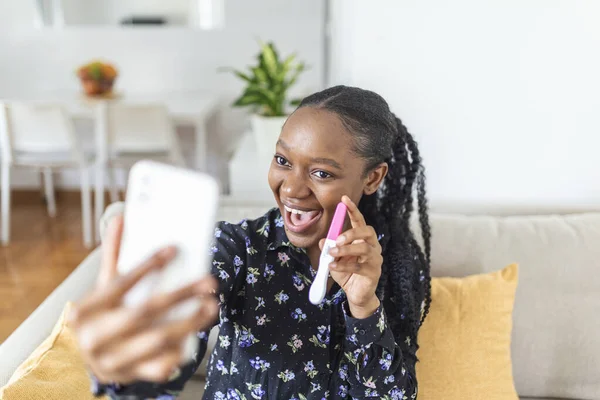 Happy, pregnant African woman is showing her pregnancy test and taking selfie making video call. Happy woman taking photo of pregnancy test with mobile phone and posting picture on social media.