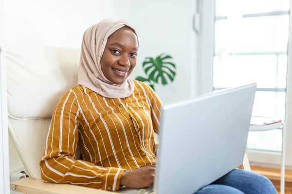 Happy pretty Muslim African woman using laptop sitting on cosy sofa. Beautiful young Muslim woman is using a laptop and smiling while sitting on the sofa at home