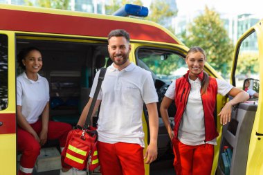 Ambulansın yanında üç sağlık görevlisi var. Genç kadın ve erkek gülümsüyor. Doktor bir tıbbi travma çantası taşıyor..
