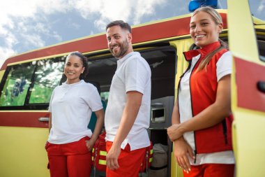 Ambulans aracının önünde portatif ekipman taşıyan üç çok ırklı sağlık görevlisi.