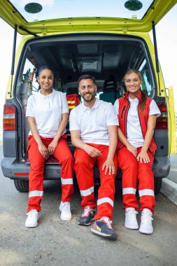 Kapıları açık bir ambulansın yanında duran çok ırklı sağlık görevlileri. İş arkadaşları tıbbi travma çantası taşıyor. Kameraya gülümsüyorlar..
