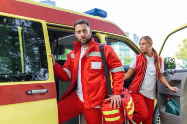 Ambulansta ilk yardım hemşiresi ve acil servis doktoru. Bir sağlık görevlisi, ambulansın arkasında duruyor, açık kapıların yanında..