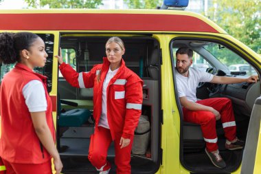 Sağlık görevlileri ve doktor ambulansın yanında. Doktor bir tıbbi travma çantası taşıyor. Üç sağlık görevlisi gülümseyerek ambulansın önünde duruyor..