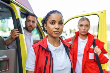 Three multiracial paramedics standing in front of ambulance vehicle, carrying portable equipment clipart