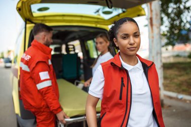 Genç Afro-Amerikan sağlık görevlisi, gülümsüyor, ambulansın arkasında duruyor, kollarını kavuşturmuş. Ambulansın yanında üç sağlık görevlisi var.