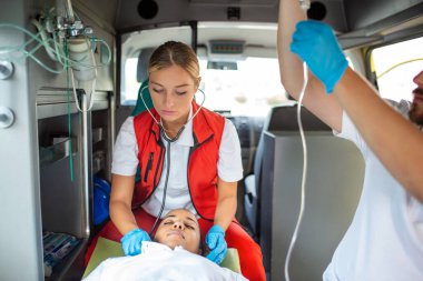 Yaralı hastayla ilgilenen sağlık görevlileri iğne yapıyor. Genç erkek sağlık görevlileri serum solüsyonu tutuyor. Kadın doktor, hastanın kalbini dinliyor.