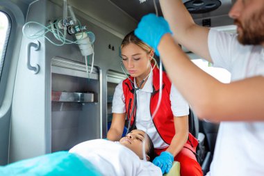Yaralı hastayla ilgilenen sağlık görevlileri iğne yapıyor. Genç erkek sağlık görevlileri serum solüsyonu tutuyor. Kadın doktor, hastanın kalbini dinliyor.