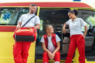Sağlık görevlileri ve doktor ambulansın yanında. Doktor bir tıbbi travma çantası taşıyor. Üç sağlık görevlisi gülümseyerek ambulansın önünde duruyor..
