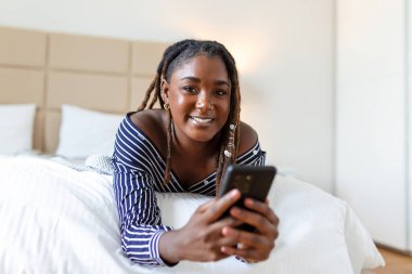 Yatakta oturan ve cep telefonu kullanan gülümseyen kadın. Gece kıyafetli Afrikalı kadın evde yatakta oturmuş cep telefonuna bakıyor..