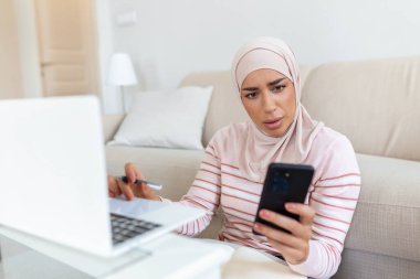 Cep telefonu ve dizüstü bilgisayar kullanarak evdeki oturma odasında online alışveriş bilgilerini arayan zarif, çekici bir Müslüman kadın. İnternet üzerinden ürün alan mutlu kadın portresi.
