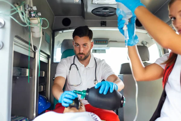 EMS Paramedics Team Provide Medical Help to Injured Patient on the Way to Healthcare Hospital. Emergency Care Assistant Using Ventilation Mask in an Ambulance. Young female nurse holding iv solution