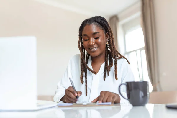 Beautiful African woman freelancer noting information for planning project doing remote job via laptop computer. woman laughing while reading email on modern laptop device .