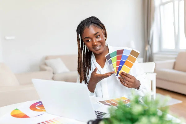 Cheerful african american lady designer having video conference with clients, sitting at desk in front of computer, holding color palettes, gesturing and smiling, copy space